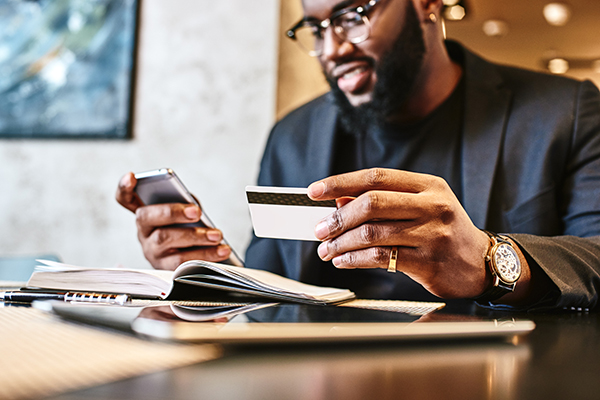 African American Male holding credit card and phone in the other with business documents and tablet on the table.