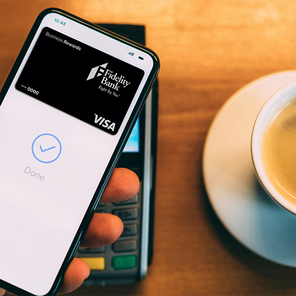 Close up of hand holding a mobile phone and paying with their digital wallet. Fidelity Bank Business Rewards credit card is on the phone screen. A coffee cup is sitting on a table next to the payment terminal.