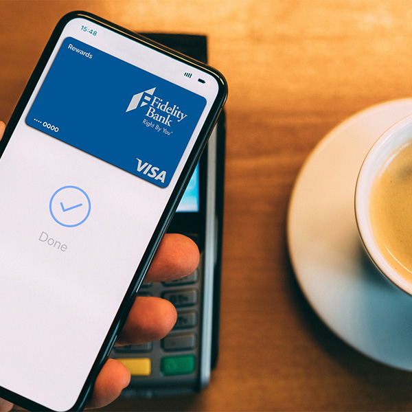 Close up of hand holding a mobile phone and paying with their digital wallet. Fidelity Bank Rewards credit card is on the phone screen. A coffee cup is sitting on a table next to the payment terminal.